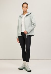 Ljusgrön vattentät jacka med huva, dragkedjestängning och mönstrade paneler, kombinerad med svarta leggings och ljusgröna träningsskor.