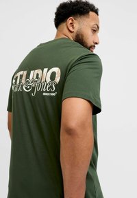 Camiseta verde de algodón con un gran estampado texturizado "STUDIO Jack & Jones" en colores claros en la espalda, cuello redondo y mangas cortas.