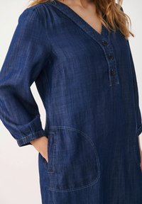 Vestido túnica de mezclilla en azul oscuro, con escote en V, mangas tres cuartos con puños elásticos y bolsillos laterales con bordes redondeados.