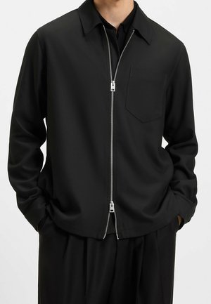 Mann trägt ein schwarzes langärmeliges Hemd mit Kragen, vorne mit Doppelreißverschluss und Brusttasche, Hände in den Taschen.