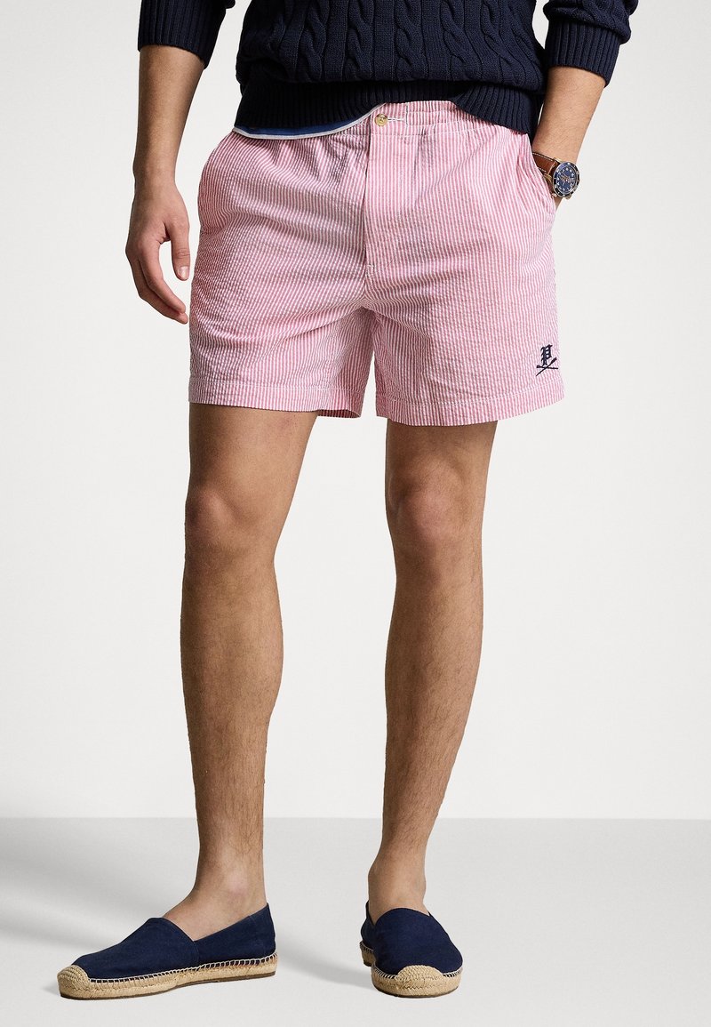 Homme portant un short rayé rose et blanc avec un logo brodé, un pull marine, une montre marron et des espadrilles bleu marine, debout devant un fond blanc.