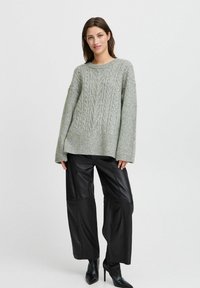 Hellgrauer Kabelstrickpullover mit Oversized-Passform, strukturiertem Muster und überschnittenen Schultern, kombiniert mit schwarzen, weit geschnittenen Lederhosen.