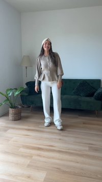 Cardigã bege tricotado com botões, mangas bufantes; calças brancas; usando uma faixa de cabelo branca; sofá verde e planta ao fundo.