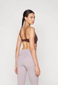 Καφέ αθλητικό σουτιέν με ρυθμιζόμενες τιράντες και κλείσιμο στην πλάτη, συνδυασμένο με ανοιχτό μωβ ψηλόμεσα κολάν, λεία υφή, μινιμαλιστικός σχεδιασμός.