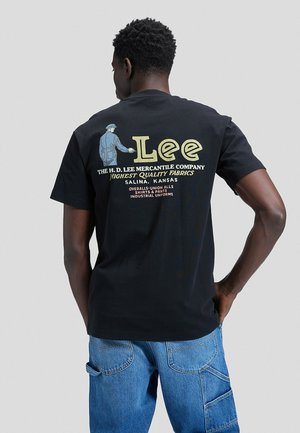 Uomo che indossa una t-shirt Lee nera con logo vintage e testo sul retro, jeans blu, in piedi con una mano sui fianchi davanti a uno sfondo grigio uniforme.