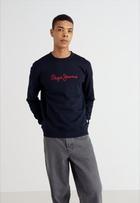Sudadera azul marina de algodón pesado, con un logo bordado en rojo de "Pepe Jeans" en el pecho. Acompañada de pantalones grises.