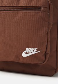 Bruine polyester rugzak met een gestructureerd oppervlak, een ritsvak en een wit geborduurd Nike-logo aan de voorkant.