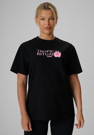 Blond kobieta w czarnej koszulce z różowym napisem "Tropical Ritual" i różowym kwiatem hibiskusa, stojąca na jednolitym szarym tle.