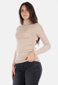 Maglione a collo alto beige a coste con maniche lunghe, caratterizzato da bordi ondulati. Tessuto morbido con un delicato luccichio, abbinato a jeans di denim scuri.