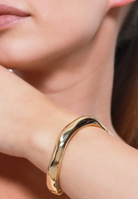 Bracciale rigido in oro con un design liscio e ondulato. Superficie riflettente, senza decorazioni, avvolge il polso comodamente.
