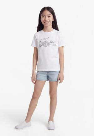 Ragazza sorridente con capelli lunghi e scuri che indossa una t-shirt bianca con grafica, pantaloncini di jeans chiari e scarpe slip-on bianche, in piedi su sfondo bianco.