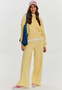 Žlutá mikina s nápisem, odpovídající široké kalhoty a růžové tenisky. Doplňky zahrnují růžovou kšiltovku a modrou kabelku přes rameno.