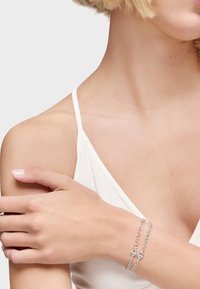 Bracciale a catena d'argento con maglie intrecciate e chiusura, indossato su una mano appoggiata su un braccio leggermente abbronzato. Design semplice, superficie liscia.