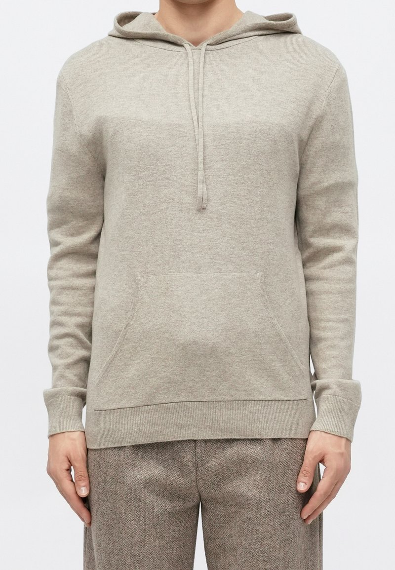 Hoodie beige en maille avec poche avant, cordons et poignets côtelés, porté avec un pantalon marron texturé.