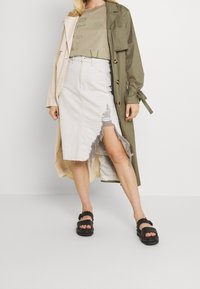 Woman wearing beige distressed denim skirt with front slit, olive green graphic t-shirt, two-toned trench coat, and black sandals.