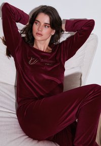 Burgundska žametna loungewear komplet, ki vključuje dolg rokav zgornji del s napisom "sledi svojim sanjam" in usklajene hlače, mehka tekstura, sproščen kroj.