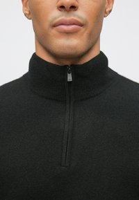 Juodas, su užtrauktuku, aukštu apykaklės megztinis, pagamintas iš tekstūruoto trikotažo. Turi mažą užtrauktuko laikiklį ties kaklu ir pasižymi prigludusia forma.