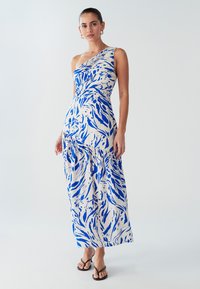 Robe maxi asymétrique bleu à motifs sur tissu crème, avec un corsage ajusté et une jupe fluide. Portée avec des tongs noires.