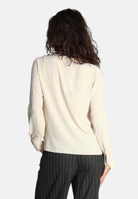 Blusa beige a maniche lunghe realizzata in tessuto leggero. Presenta polsini con bottoni e una vestibilità morbida, abbinata a pantaloni scuri a righe sottili.