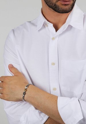 Bracciale in argento e tonalità oro caratterizzato da anelli metallici intrecciati. Indossato su un polso, abbinato a una camicia bianca con bottoni.