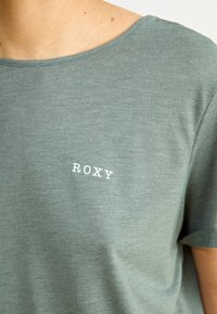 Olivovo zelené tričko s krátkym rukávom z jemnej látky s okrúhlym výstrihom. Na ľavej strane hrudníka sa nachádza biele logo "ROXY".