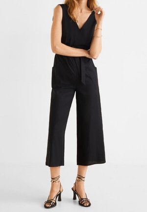 Woman wearing a sleeveless black jumpsuit with wide legs and a waist tie, paired with black strappy high-heeled sandals.