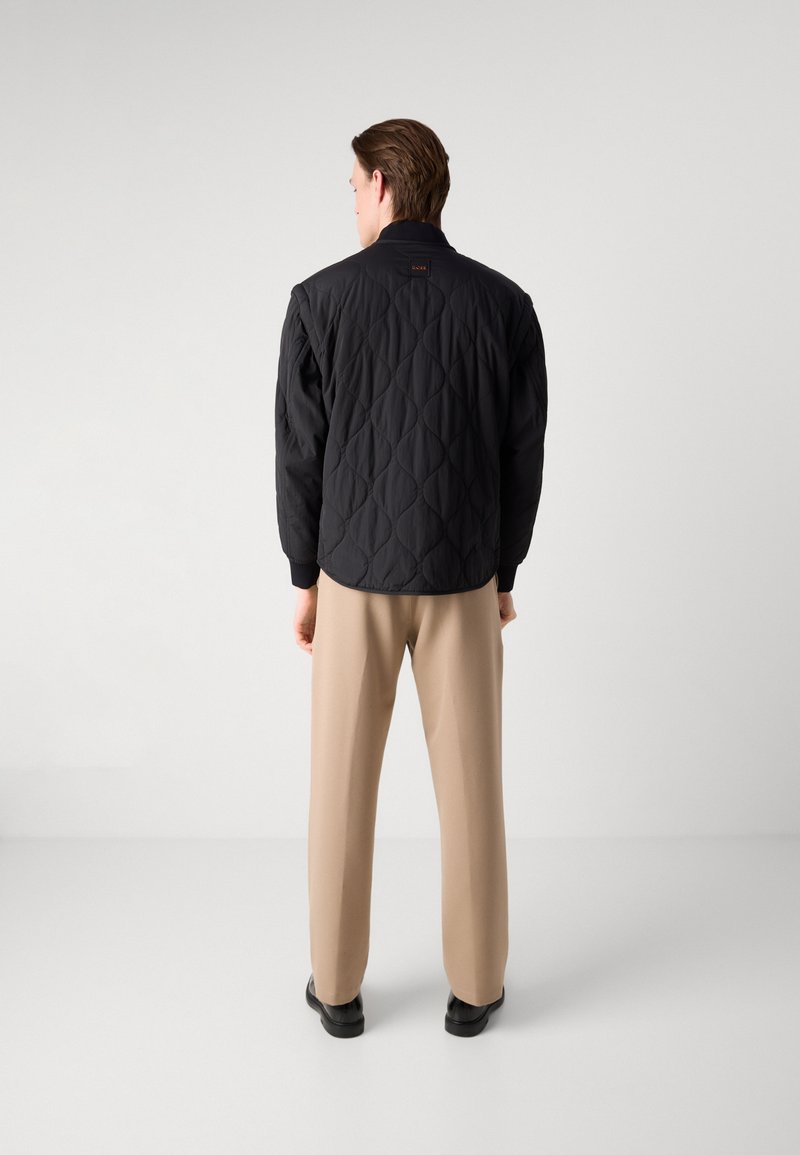 Schwarze gesteppte Jacke mit Stehkragen, strukturiertem Rautenmuster und Druckknopfverschluss, kombiniert mit hellbeigen Hosen.