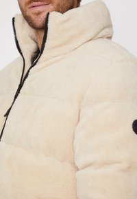 Chaqueta de forro polar beige con un alto cuello, cierre con cremallera en el frente y detalles en negro, que muestra una textura suave y un patrón acolchado.
