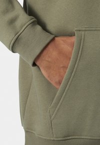 Sudadera verde oliva de algodón suave, con un bolsillo frontal y puños acanalados. Detalles de costura y textura visibles, que muestran durabilidad.