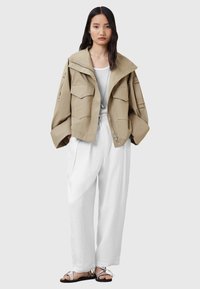 Giacca beige a croppata con ampie tasche frontali, maniche larghe e colletto alto, abbinata a pantaloni bianchi larghi e sandali flat.
