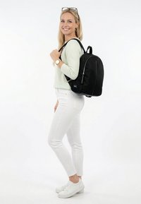 Žena v lehkém svetru a bílých kalhotách stojí usmívající se, přičemž přes jedno rameno nese černý batoh na jednobarevném bílém pozadí.