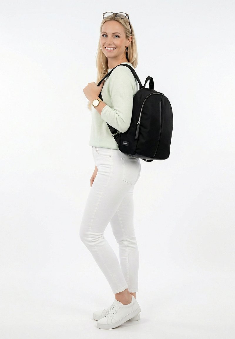 Žena v lehkém svetru a bílých kalhotách stojí usmívající se, přičemž přes jedno rameno nese černý batoh na jednobarevném bílém pozadí.