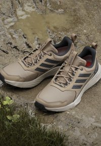 Beige athletic shoes with black accents, mesh upper, rubber sole, and textured design. Features laces and a pull tab on the heel.