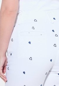 Pantalones blancos con un patrón repetido de pequeños corazones azules, con un bolsillo trasero estándar y lazos para el cinturón, confeccionados en una tela ligera.