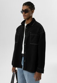 Zwarte spijkerblouse met witte stiksel, enkele borstzak, gedragen over een wit T-shirt. Lichte spijkerbroek zichtbaar, tas in de hand.