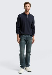 Marineblaues Langarm-Polohemd mit einer Drei-Knopf-Leiste, kombiniert mit ausgewaschenen blauen Jeans und weißen Sneakers. Einfaches, lässiges Design.