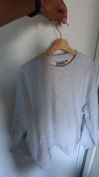 Hand holding wooden hanger with light gray Carhartt sweatshirt against white door background.