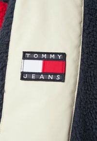 Tommy Jeans-lap med sorte, hvide og røde sektioner på en cremefarvet baggrund, med tekstureret marineblå stof ved siden af.