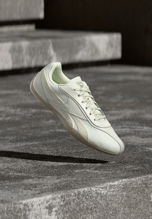 Λευκό χαμηλό sneaker με κορδόνια που αιωρείται πάνω από ένα γκρι σκυρόδεμα σκαλοπάτι σε αστικό υπαίθριο περιβάλλον.