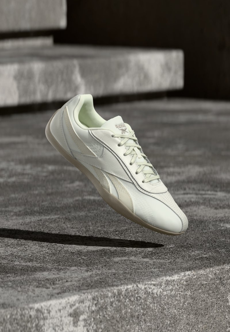 Λευκό χαμηλό sneaker με κορδόνια που αιωρείται πάνω από ένα γκρι σκυρόδεμα σκαλοπάτι σε αστικό υπαίθριο περιβάλλον.