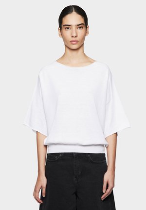 Femme aux cheveux foncés attachés, portant un haut blanc ample à manches courtes et un pantalon noir taille haute, debout devant un fond uni.
