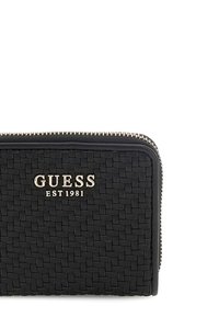 Portafoglio in pelle intrecciata nera con zip dorata e logo che recita "GUESS EST 1981." Presenta una forma rettangolare compatta e una superficie testurizzata.
