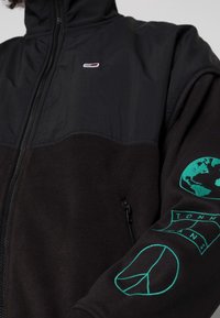 Černá fleecová bunda se zipem vpředu, s kontrastním černým nylonovým horním dílem a zelenou vyšitou grafikou na rukávu.