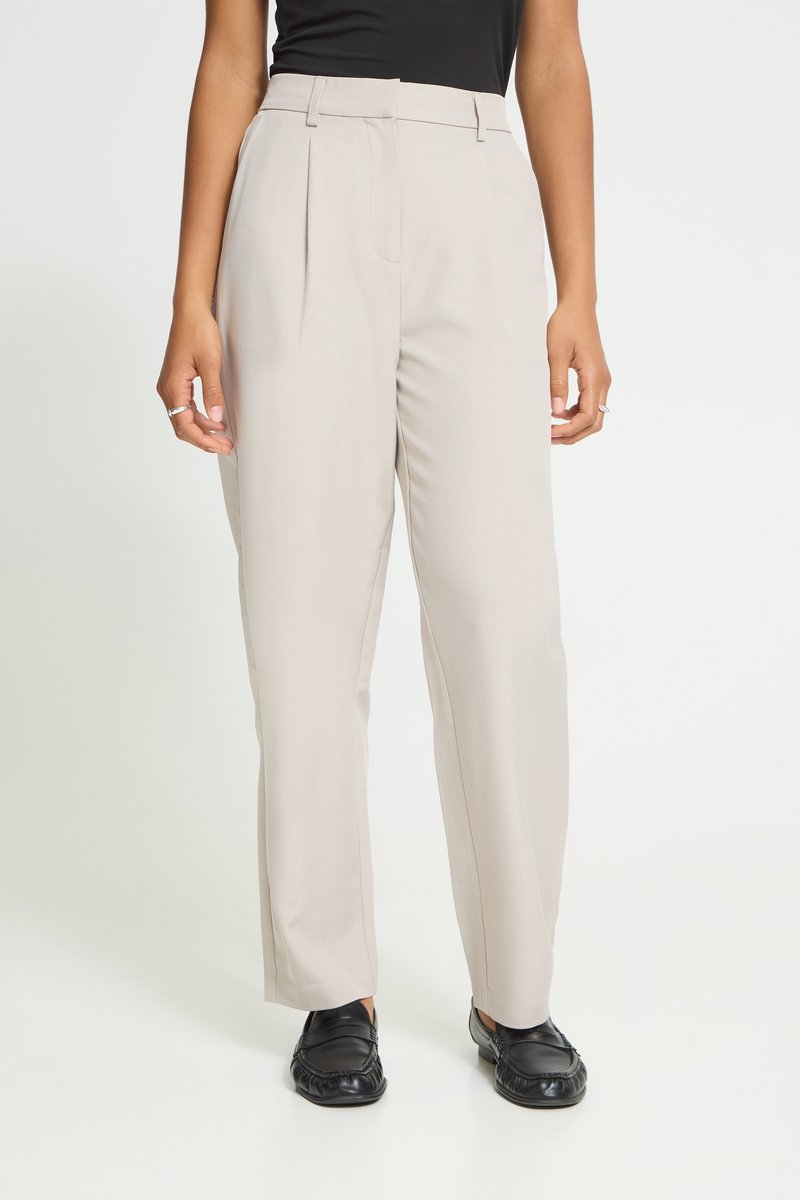 Personne portant un pantalon beige taille haute à jambes larges, un haut noir et des mocassins noirs, debout sur un fond blanc.