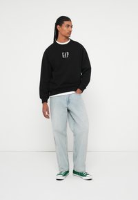 Svart genser med "GAP"-logo, hvit rund hals, lyseblå løstsittende jeans og grønne joggesko. Enkel, avslappet design.