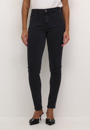 Femme portant un jean skinny noir taille haute et des mules à talons noires à bout pointu avec un détail boucle argentée, debout contre un mur beige.