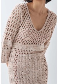 Robe en maille beige clair avec un col en V profond, un motif ajouré et des manches trois-quarts aux détails texturés variés.