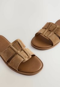 Svijetlosmeđi zamakani sandali s širokim dvostrukim remenom koji sadrži geometrijski naglasak. Potplat je od glatke smeđe kože, što pruža minimalistički dizajn.