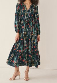 Robe maxi à imprimé floral en vert foncé, avec des manches longues, un décolleté en v, une jupe à volants et une ceinture à nouer à la taille. Associée à des talons beiges à brides.
