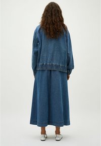 Chaqueta de denim con cintura fruncida y mangas largas, combinada con una falda larga de denim. Ambas prendas son de un tono azul medio.